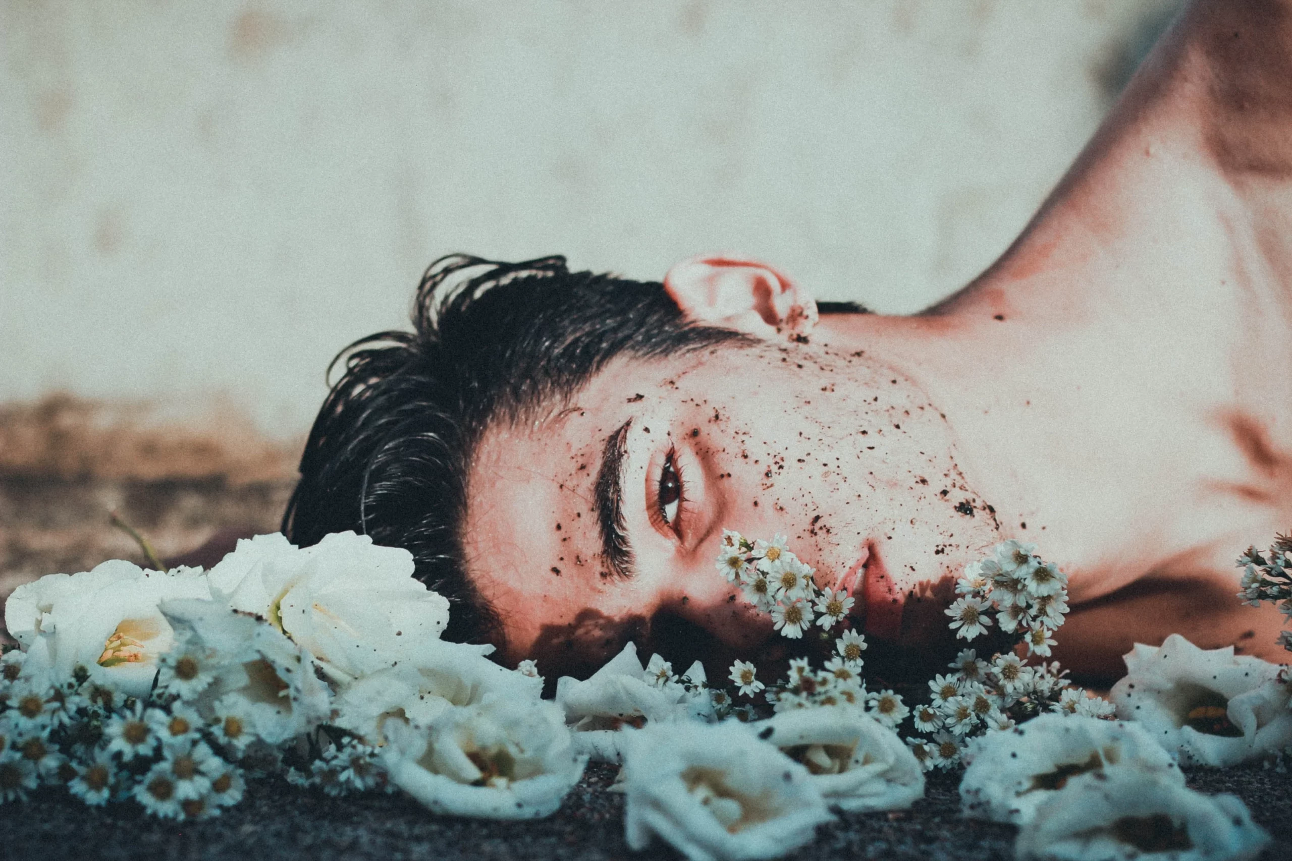 Un joven con el pecho descubierto, tumbado de lado sobre un suelo de tierra húmeda y flores blancas, con el pelo mojado y restos de tierra húmeda en la cara. Está mirando directamente al espectador.